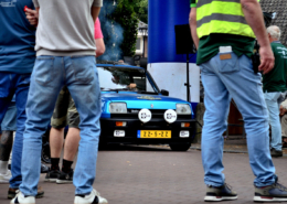 SCP_15 front left angle view of a sky blue classic Renault Turbo car in the background, spectators and crew standing on a red brick road in the foreground. vantage point through the legs of the spectators